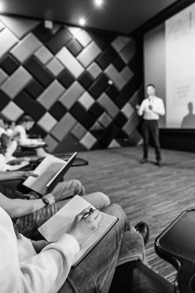 Pengumuman Dosen TA Batch II Black and white shot of students taking notes during a lecture in a modern hall.